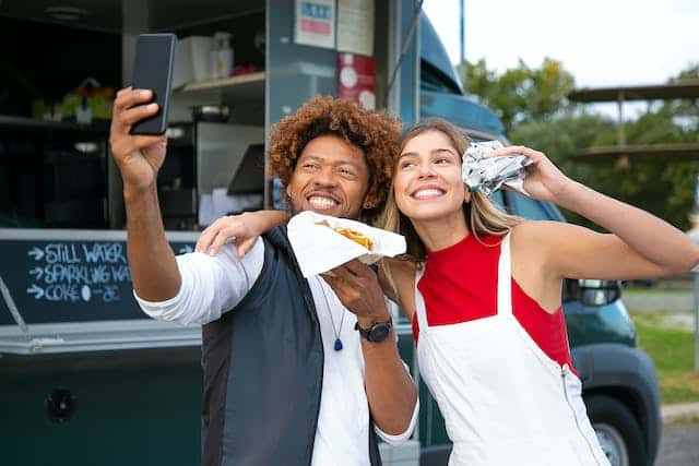 Two happy people eating food and taking a selfie in front of a food truck.